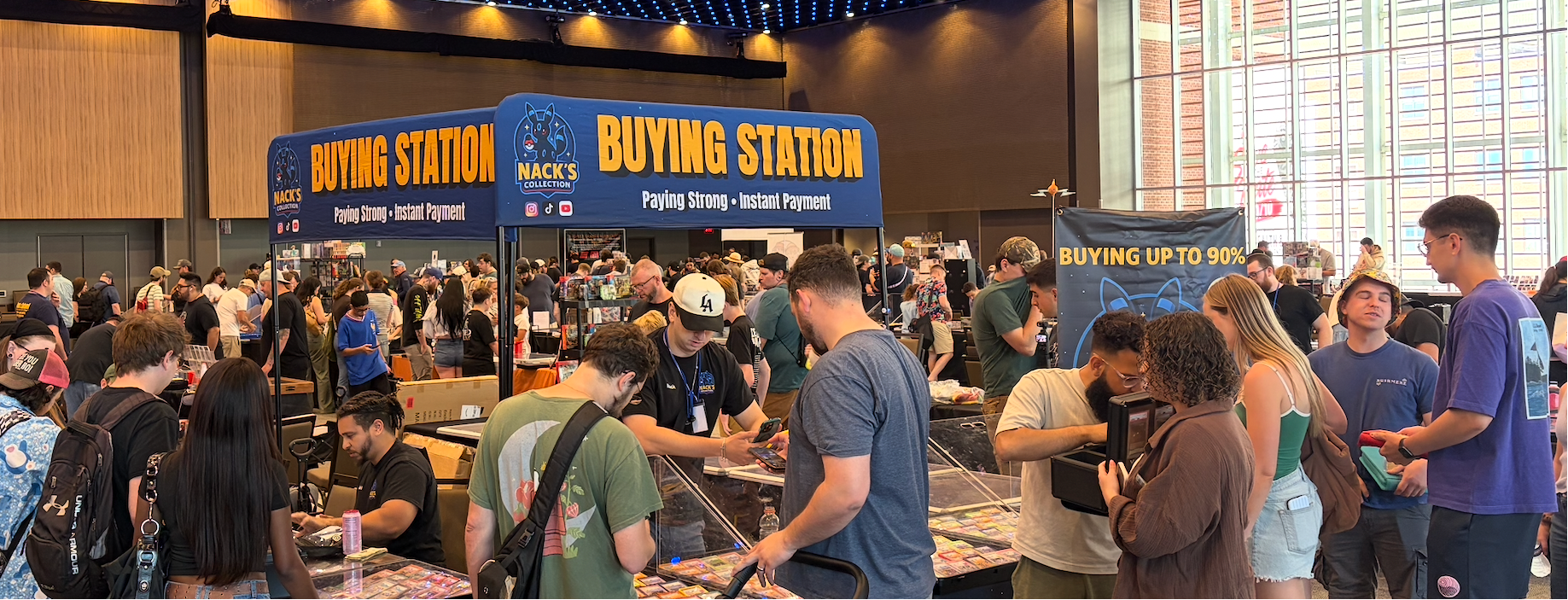 A crowded convention or fair hall with many people gathered around booths, including a prominent blue booth labeled 'BUYING STATION' with a 'Buy Again' policy sign. The scene is indoors with a high ceiling, bright windows, and many attendees.